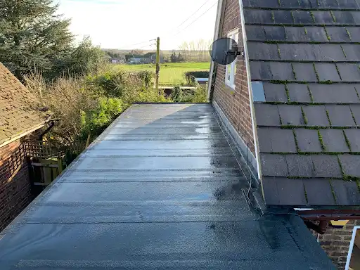 Flat black roof surface adjacent to a pitched roof with moss patches, a brick wall, and a mounted satellite dish, with trees and green fields in the background.