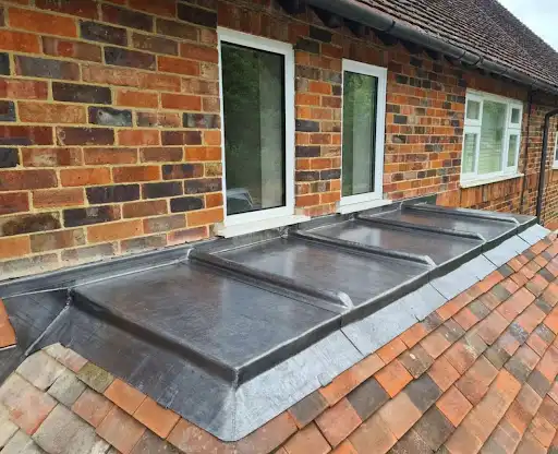 Flat metal roof section with raised seams attached to a brick house wall below two white-framed windows, adjacent to a sloped roof covered with reddish-brown shingles.