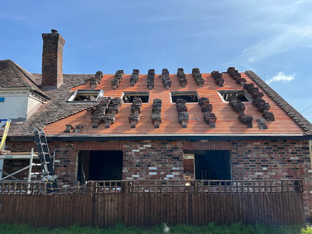 House under roof renovation with red underlayment and stacks of dark roof tiles arranged in vertical rows, with three rectangular skylights and scaffolding on the left side.