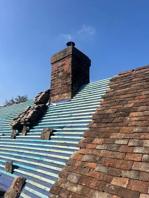 Roof with partially removed old clay tiles revealing striped underlayment, next to intact tiles and a brick chimney under blue sky.